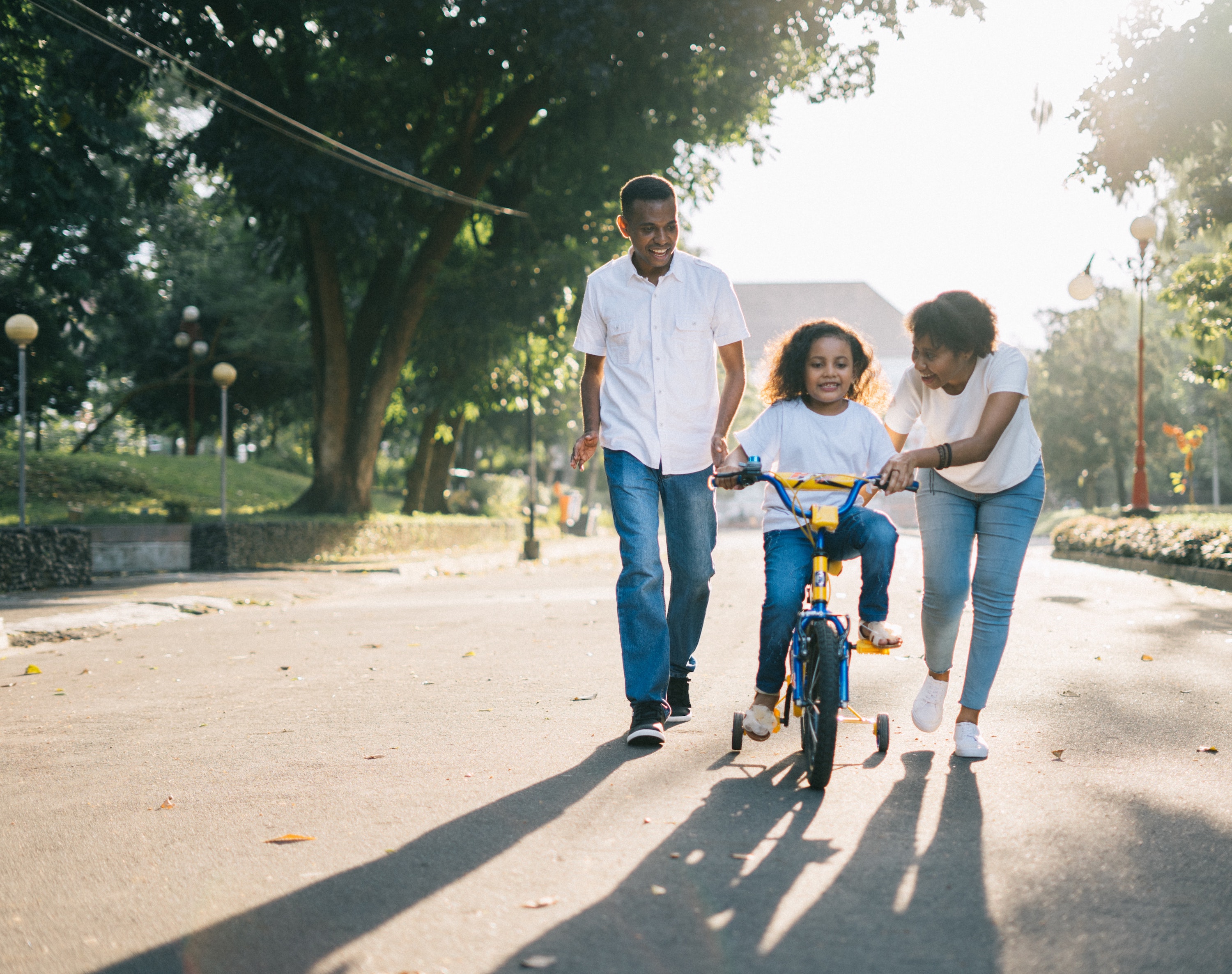 Black family of 3 all wearing blue jeans and white tops while smiling and teaching their daughter how to ride a bike