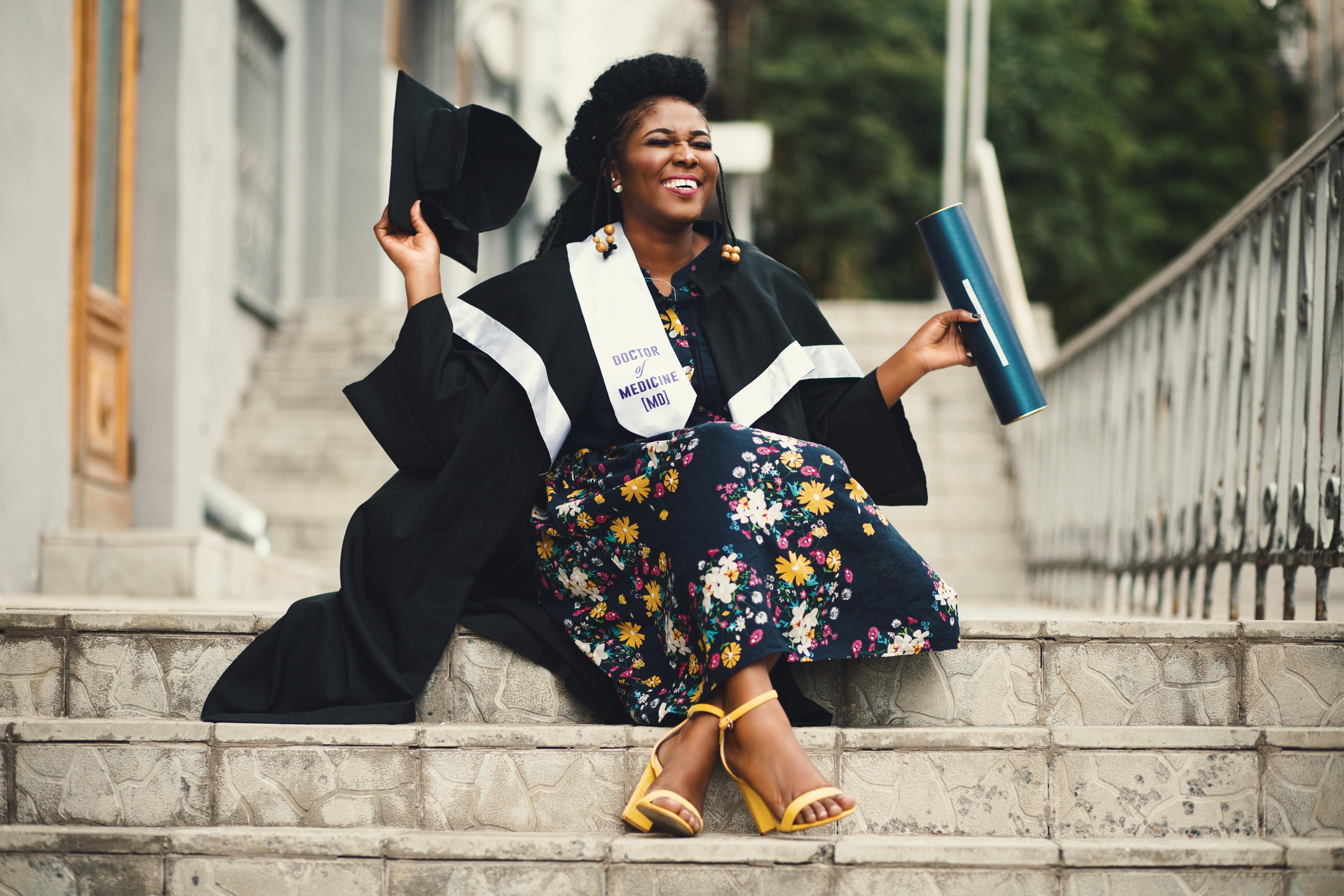 Black woman smiling while wearing her college graduation gown with her cap in hand and sitting on the steps of her college