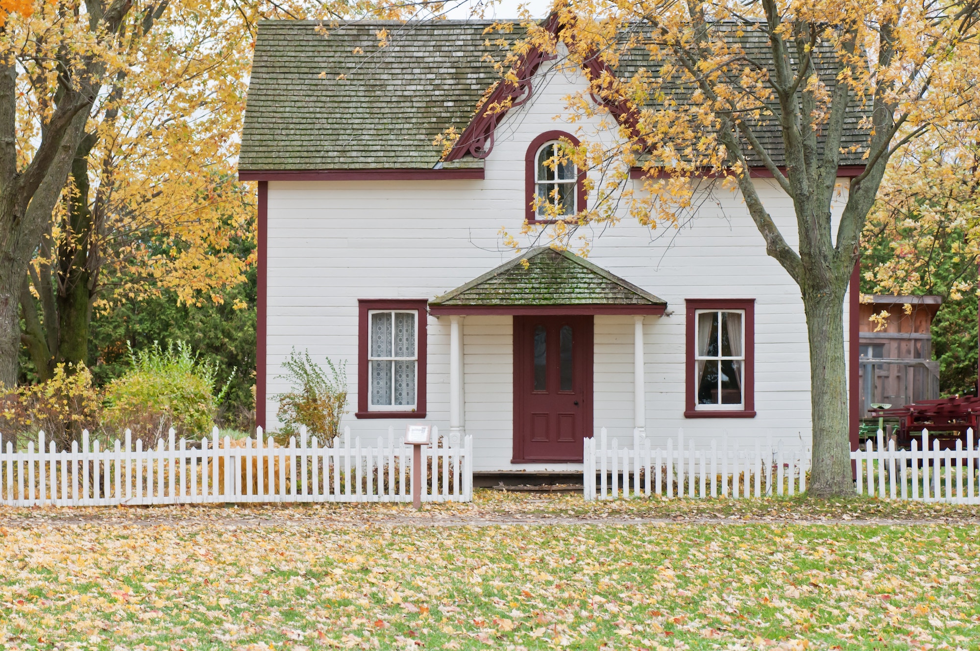Autumn leaves fall down in front of a modestly-sized white home with red trim and a green roof, surrounded by a white picket fence