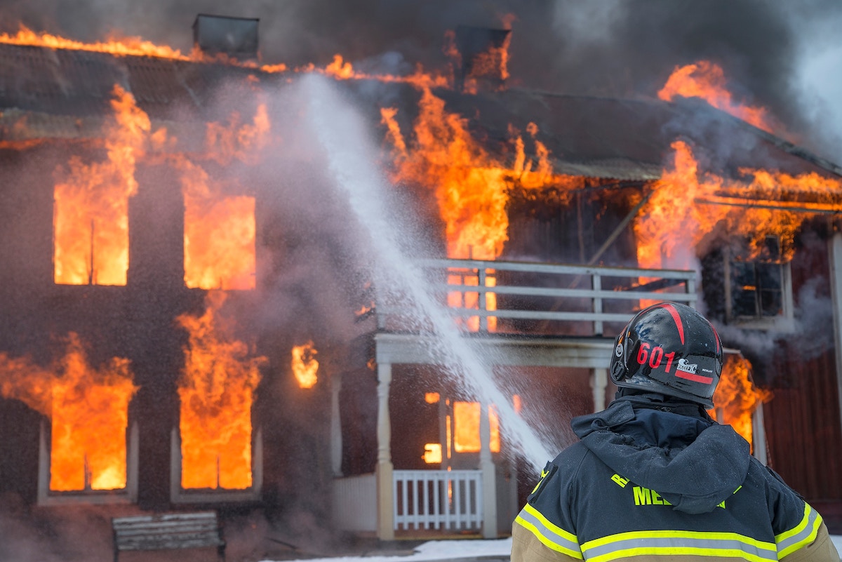 Firefighter in black and yellow suit aims his hose at a building that's up in flames