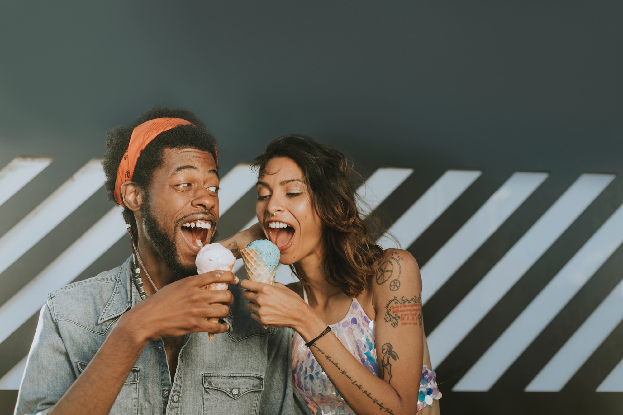 Mixed race couple makes silly faces at one another while eating ice cream in front of a funky background