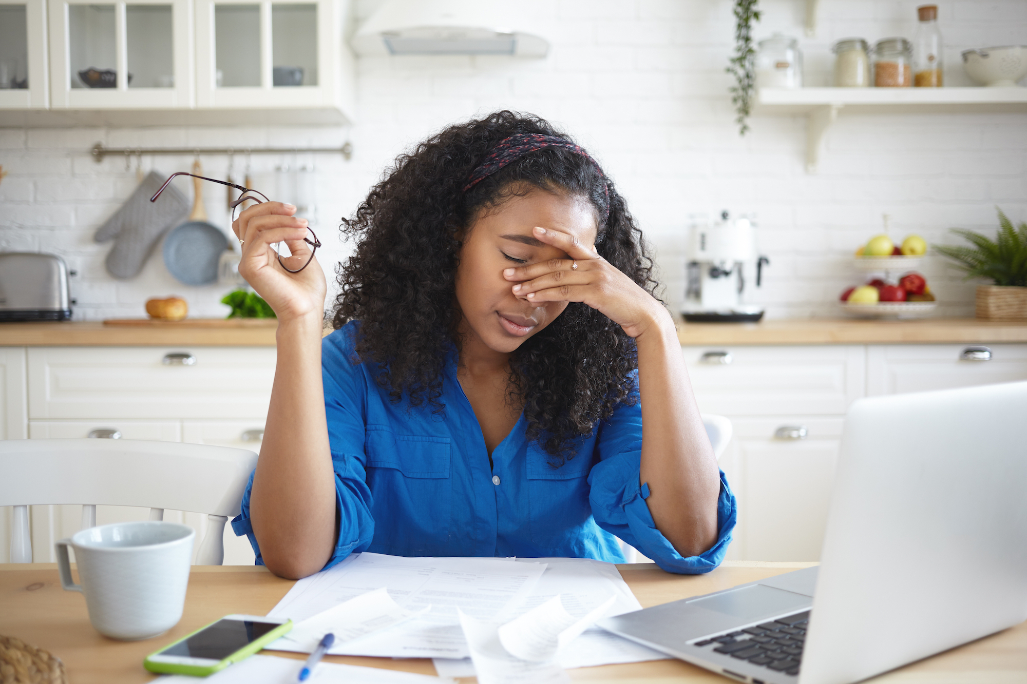 Black woman in a blue button down shirt looks stressed with her left hand on her temple and glasses in her right and sitting in her kitchen