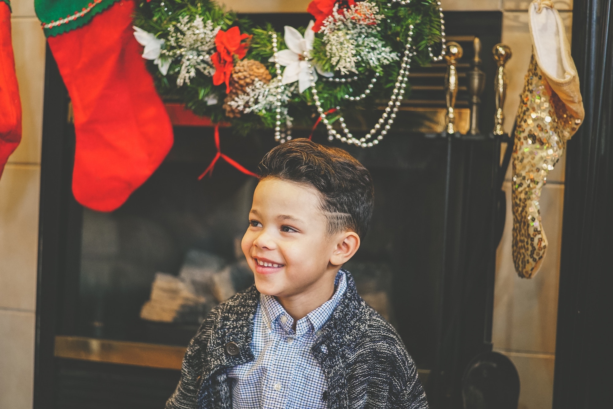 Mixed race child wearing a light blue button down shirt and dark gray cardigan sits in front of a fireplace with garland and Christmas stockings hung on it