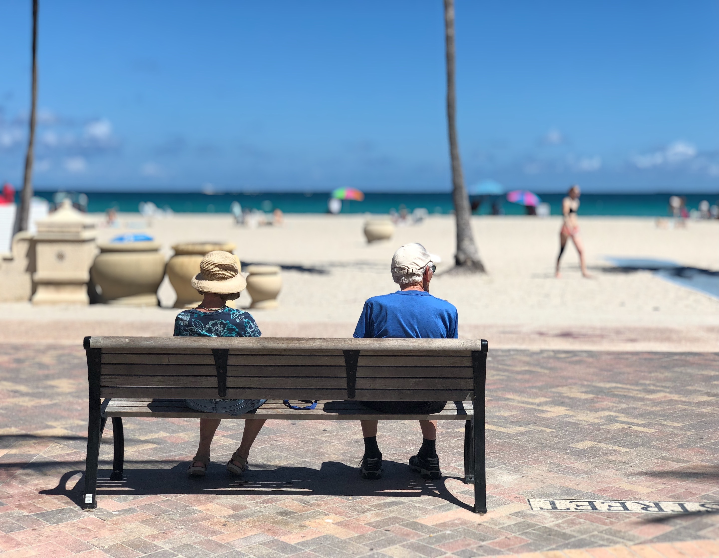 Older white couple sits on a bench on a beach sidewalk looking at the passerby