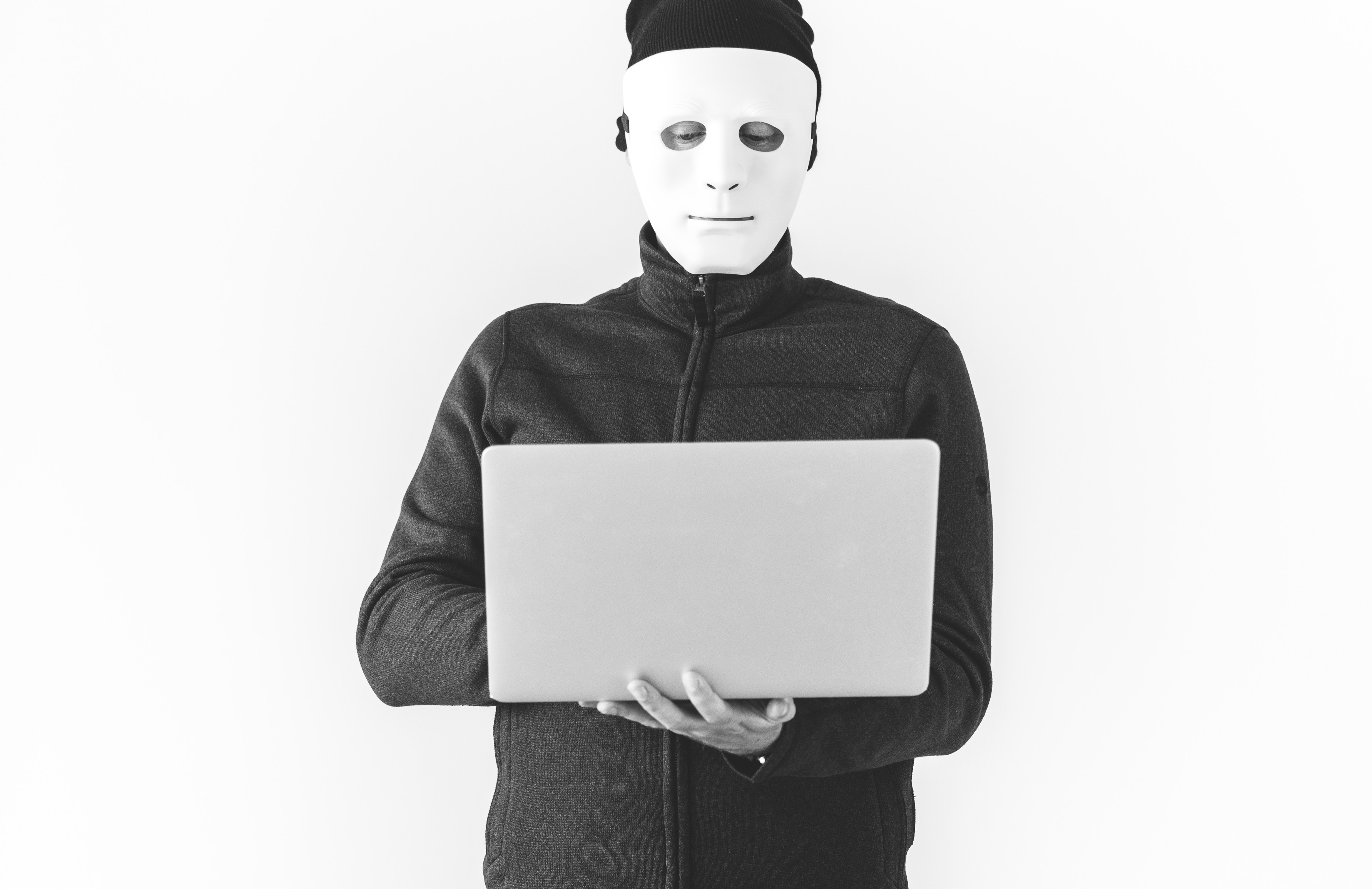Black and white photo of a man in a Jason white hockey mask wearing a black long sleeve turtleneck and black beanie suspiciously looks at a laptop