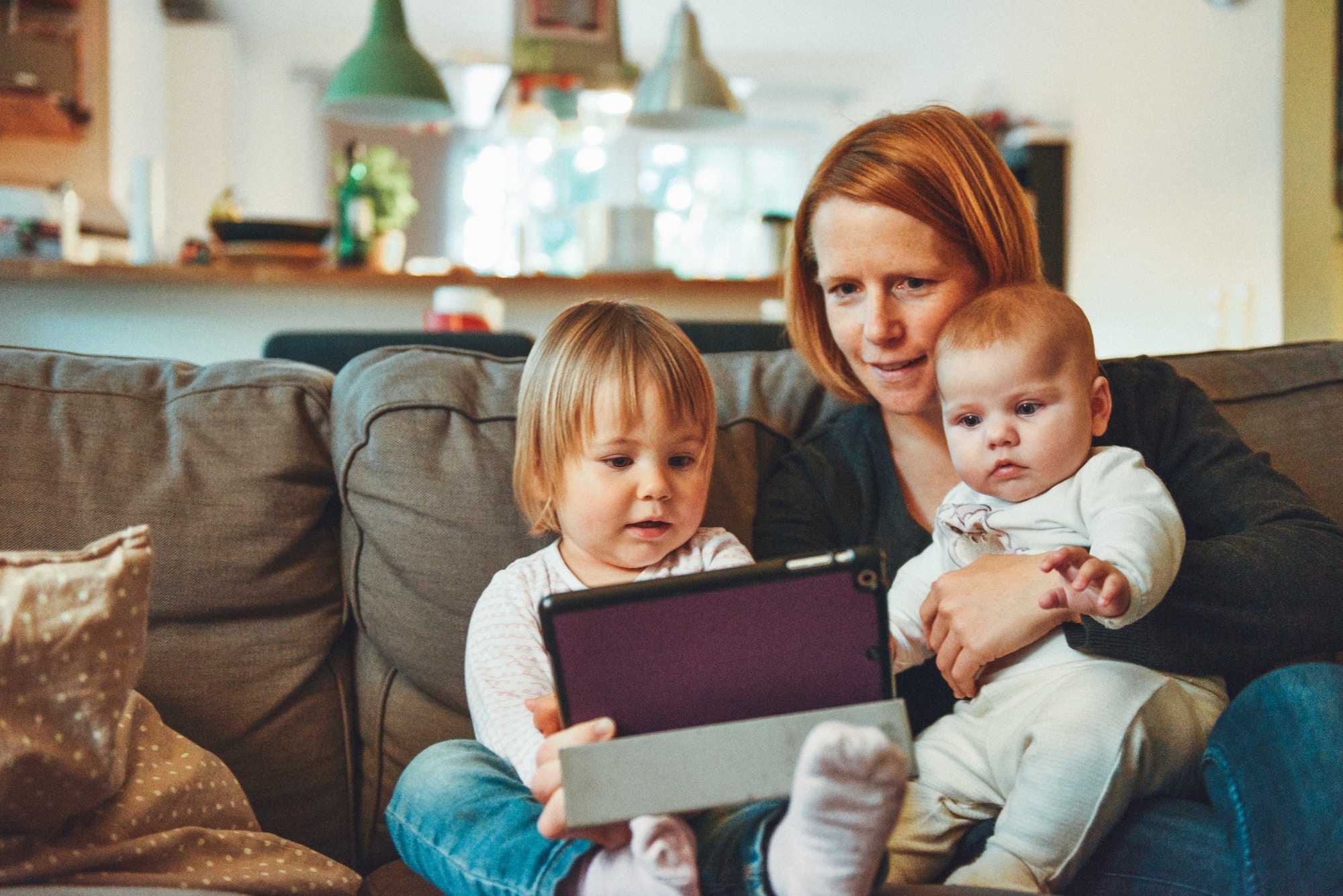 Red-headed woman sits on the couch holding her baby in her left arm while her older child sits to the right of her looking at an iPad