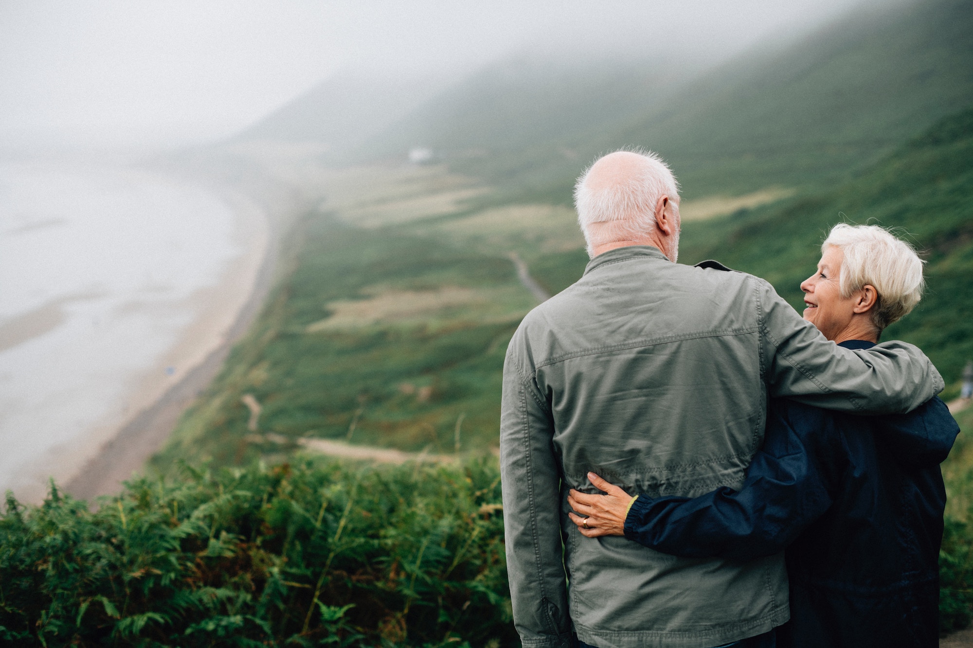 Older white couple smile at each other as they overlook the ocean while standing cliffside