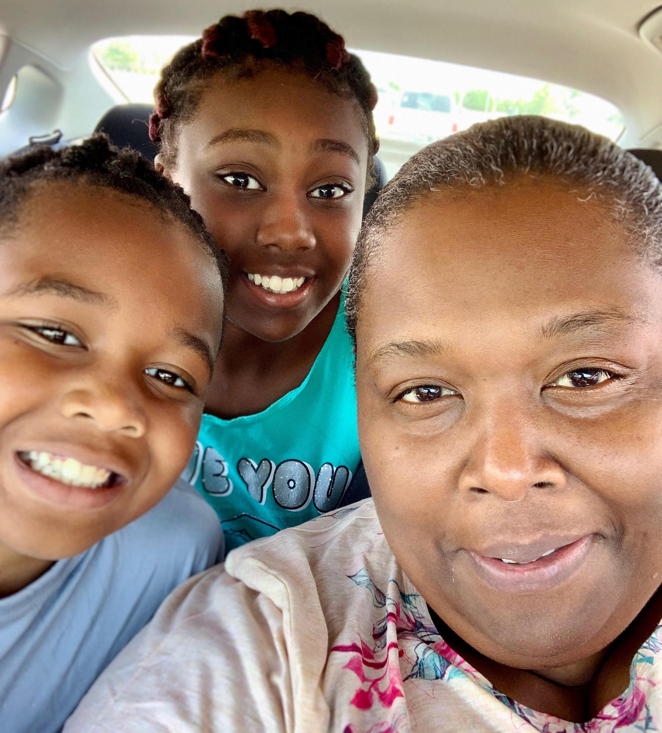 Older black woman in a tan long sleeve shirt poses with her two granddaughters in the car