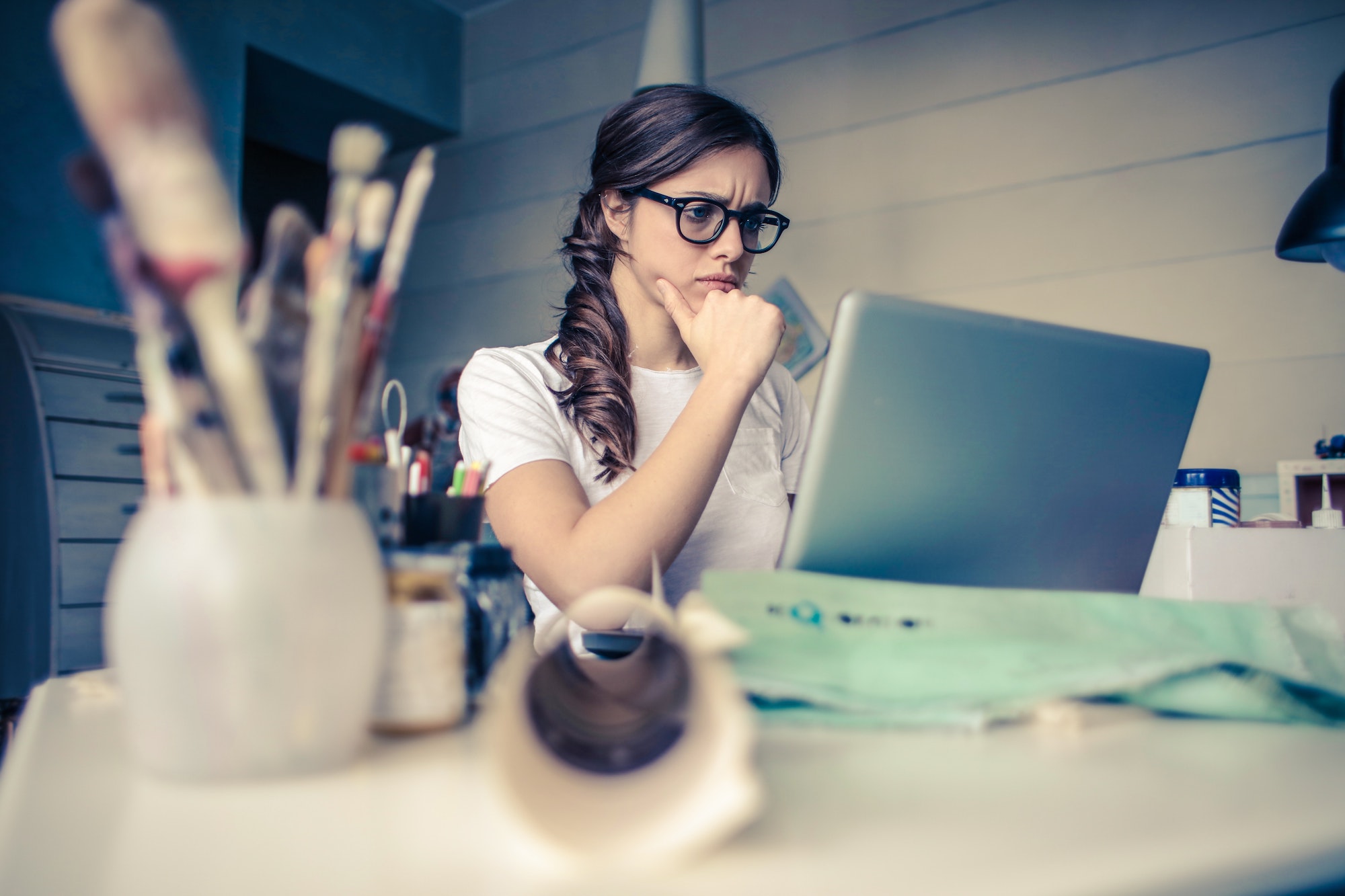 White woman in a white short sleeve t-shirt sits at her cluttered desk and looks at her laptop screen