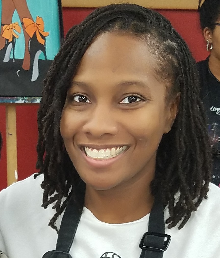 Black woman wearing a white long sleeve t-shirt and black apron while smiling in an art class