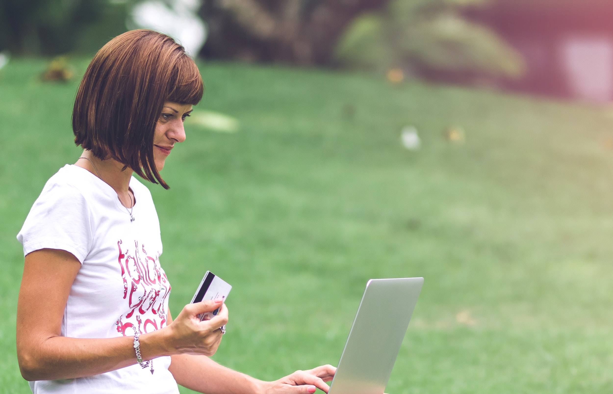 White woman with a short bob haircut and wearing a graphic white t-shirt sits in a meadow with her laptop in her lap and holding her credit card