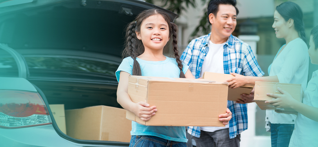 Girl in pigtails holding a box smiles for the camera while her parents are holding boxes and helping move their family into the house