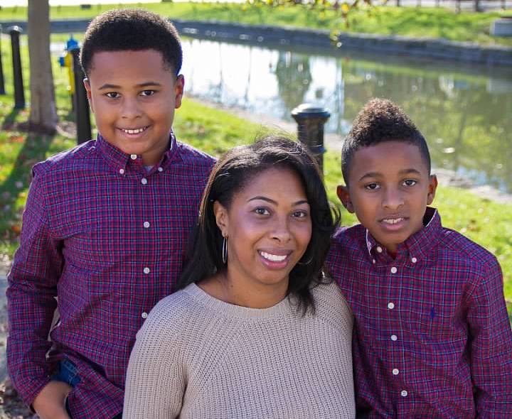 Black woman in a tan long sleeve sweater sits in between her two young sons in plaid button up shirts while their picture is being taken by a professional photographer in an outdoor setting