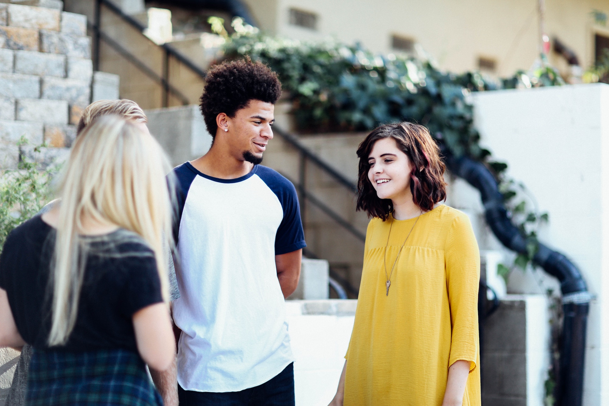 White girl wearing a yellow flowy dress and necklace smiles while talking to a black man wearing a blue and white baseball t-shirt while a blonde-haired woman has her back turned to the camera