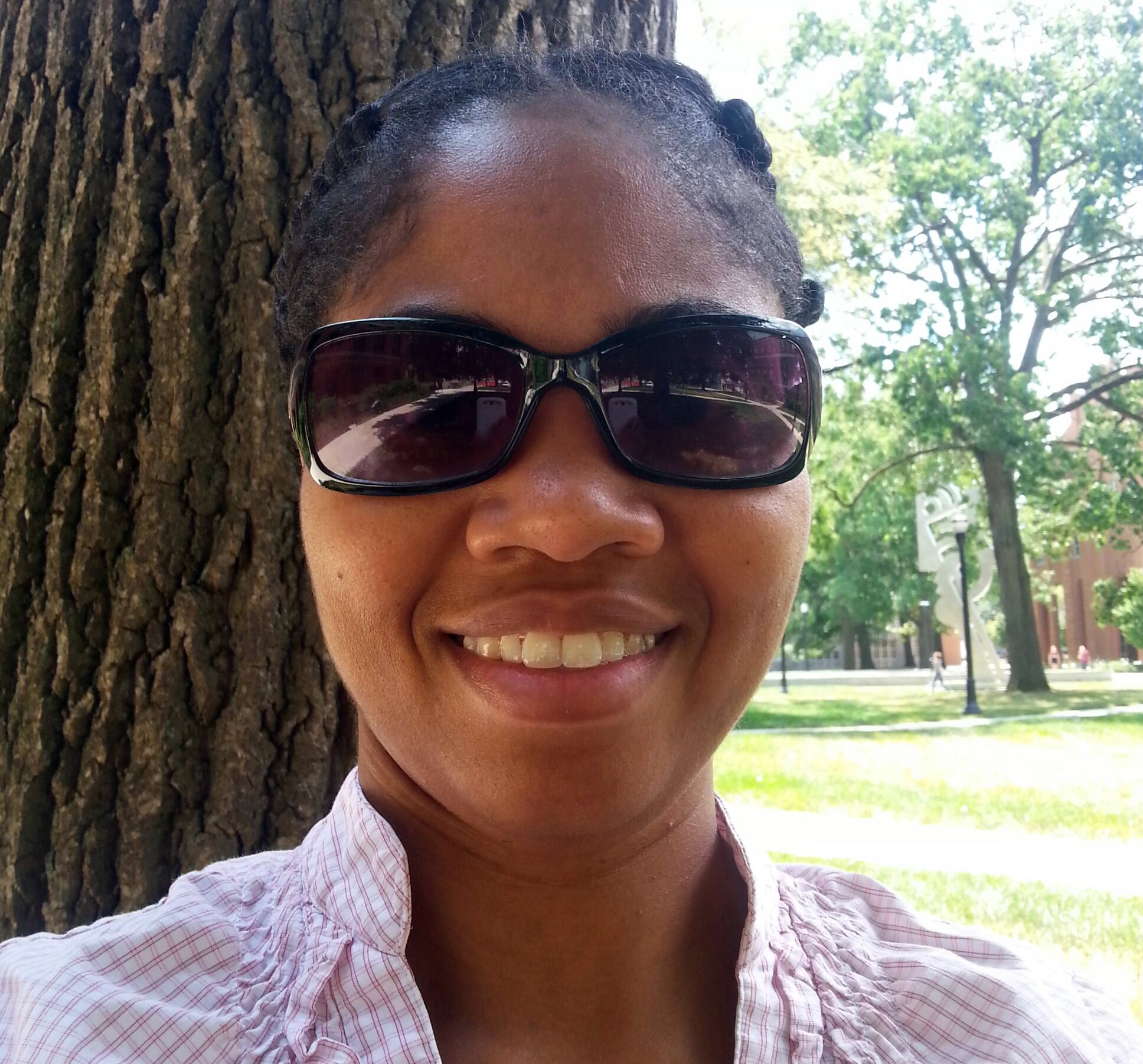 A black woman in sunglasses wears a lavender collared shirt while sitting outside by a tree