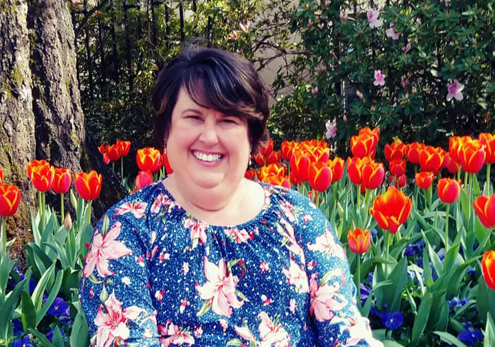White woman with a pixie haircut wears a flowery top smiles in front of a bed of tulips