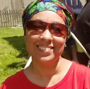 Headshot of a Black woman in sunglasses wearing a red t-shirt and bonnet smiling for the camera on a sunny day