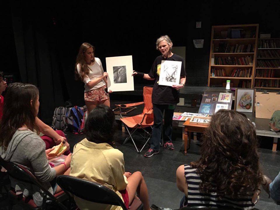 An older gray-haired woman shows her drawings to art students in a class while wearing a black graphic t-shirt, dark, jeans, and running shoes