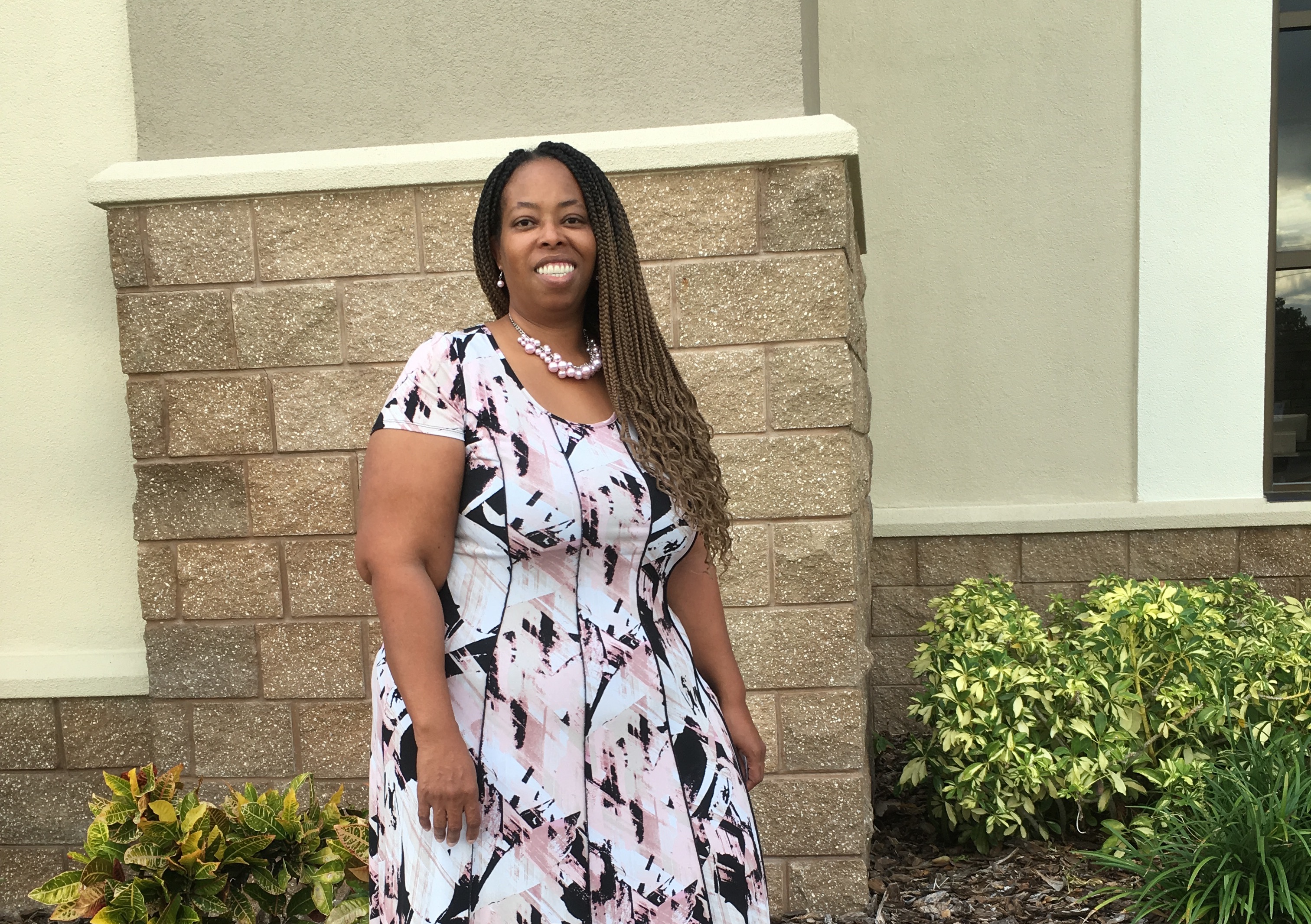 A black woman with pearl necklace wears a long white, pink, and black dress while smiling