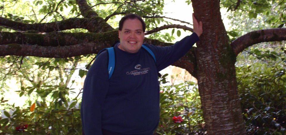 Tall man smiles while wearing a dark blue sweatshirt and carrying a light blue backpack leans one arm against a tree