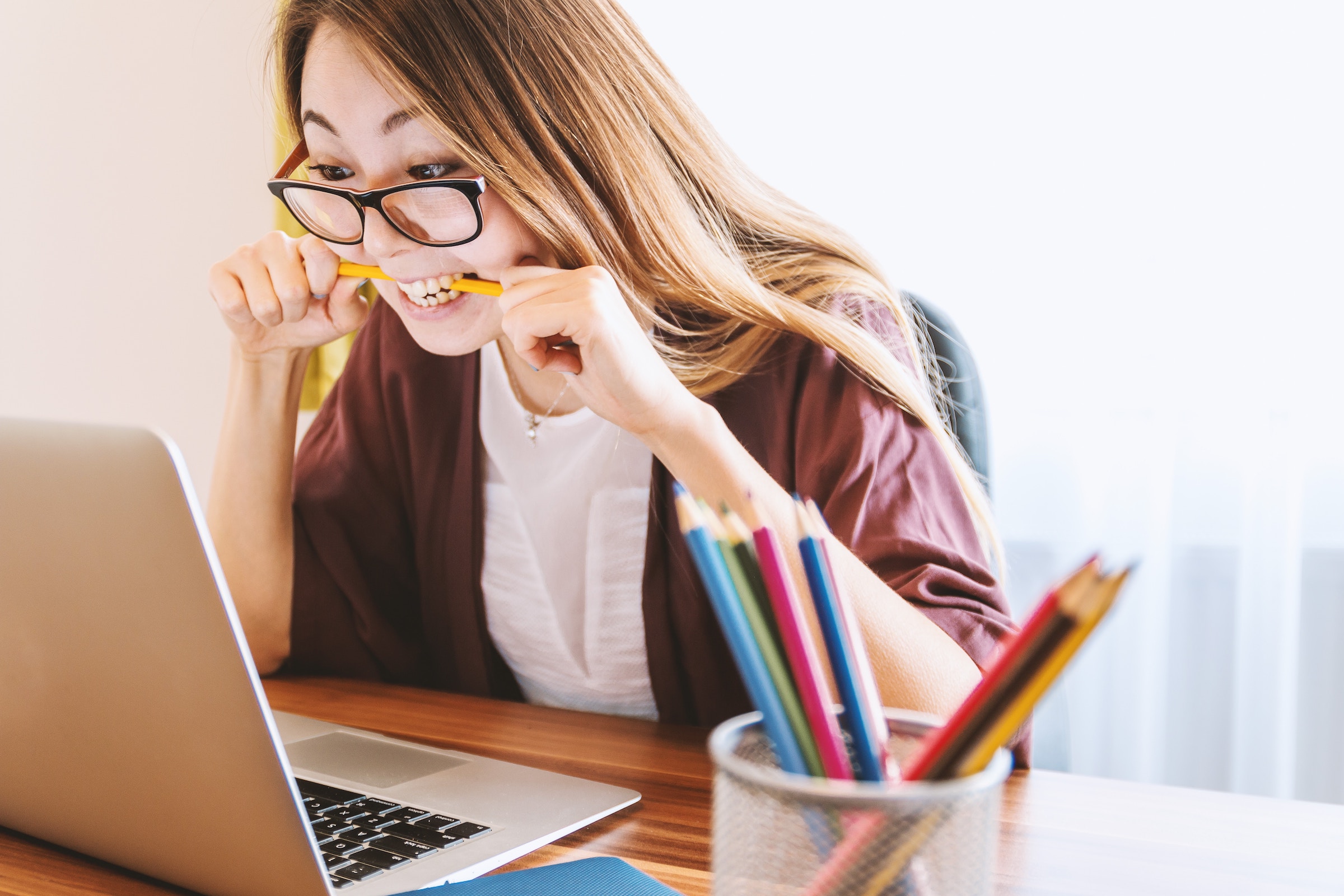 An Asian woman with glasses wearing a white shirt and maroon cardigan bites a pencil while anxiously looking at her laptop screen