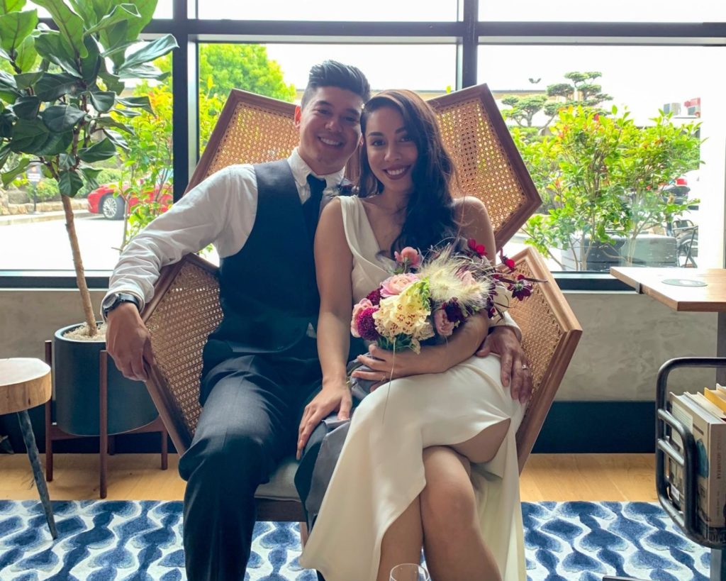 Man wearing a vest, tie, and dress pants sits next to his wife who is wearing a cream colored wedding dress and holding a bouquet of flowers on a pressed cane chair at their wedding