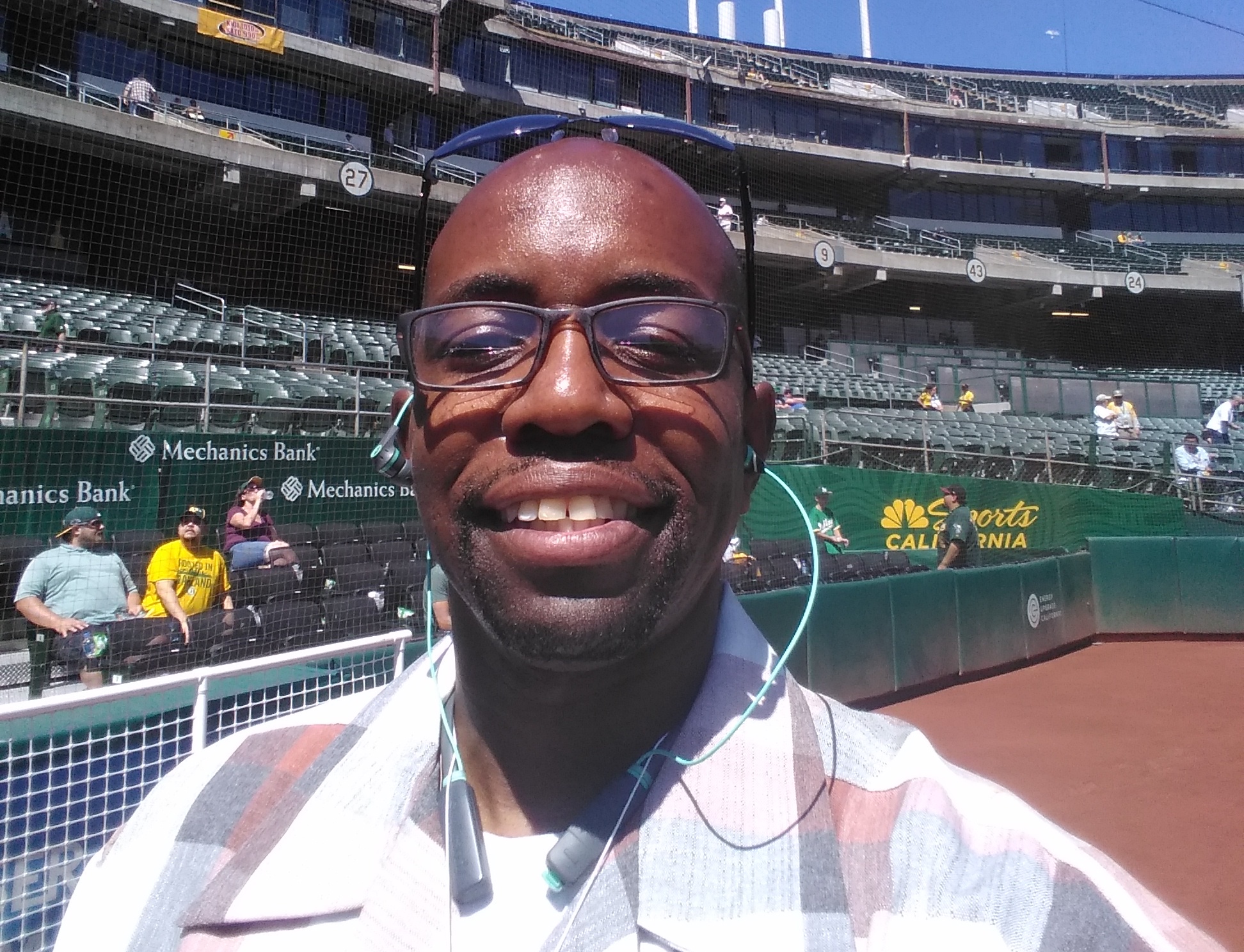 Black man with glasses wearing a checkered polo takes a selfie on the field of an empty baseball field on a sunny day