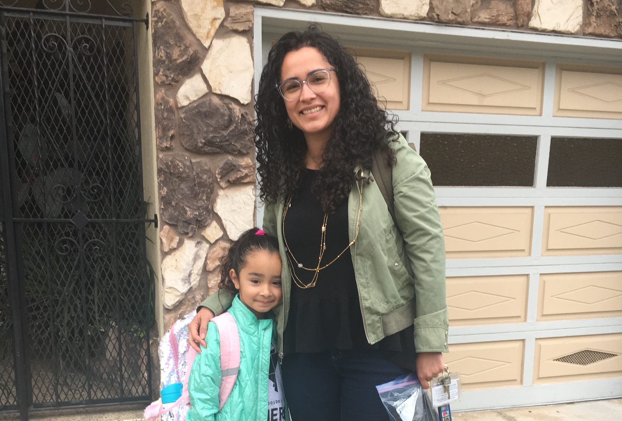 A woman with curly hair and glasses wearing a black dress and light green jacket side hugs her daughter who is wearing a teal blue jacket and backpack in front of their apartment