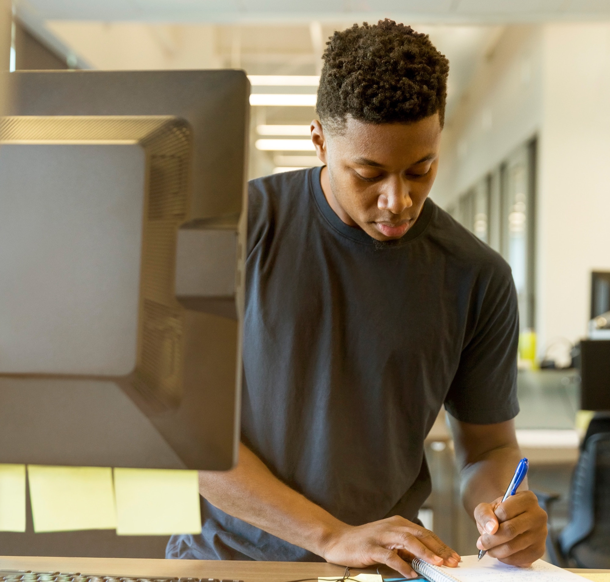 Black man wearing a black short sleeve t-shirt writes on a notebook