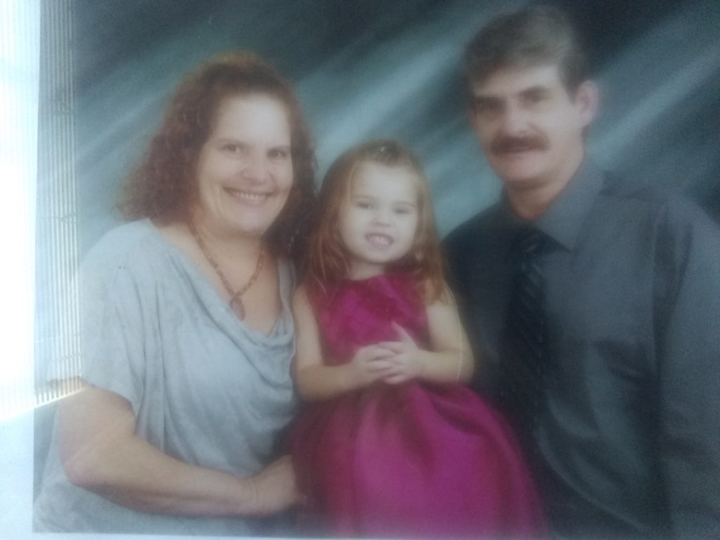 A red-headed mother, gray-haired father, and red-headed young girl pose for a picture in a professional photographer's studio