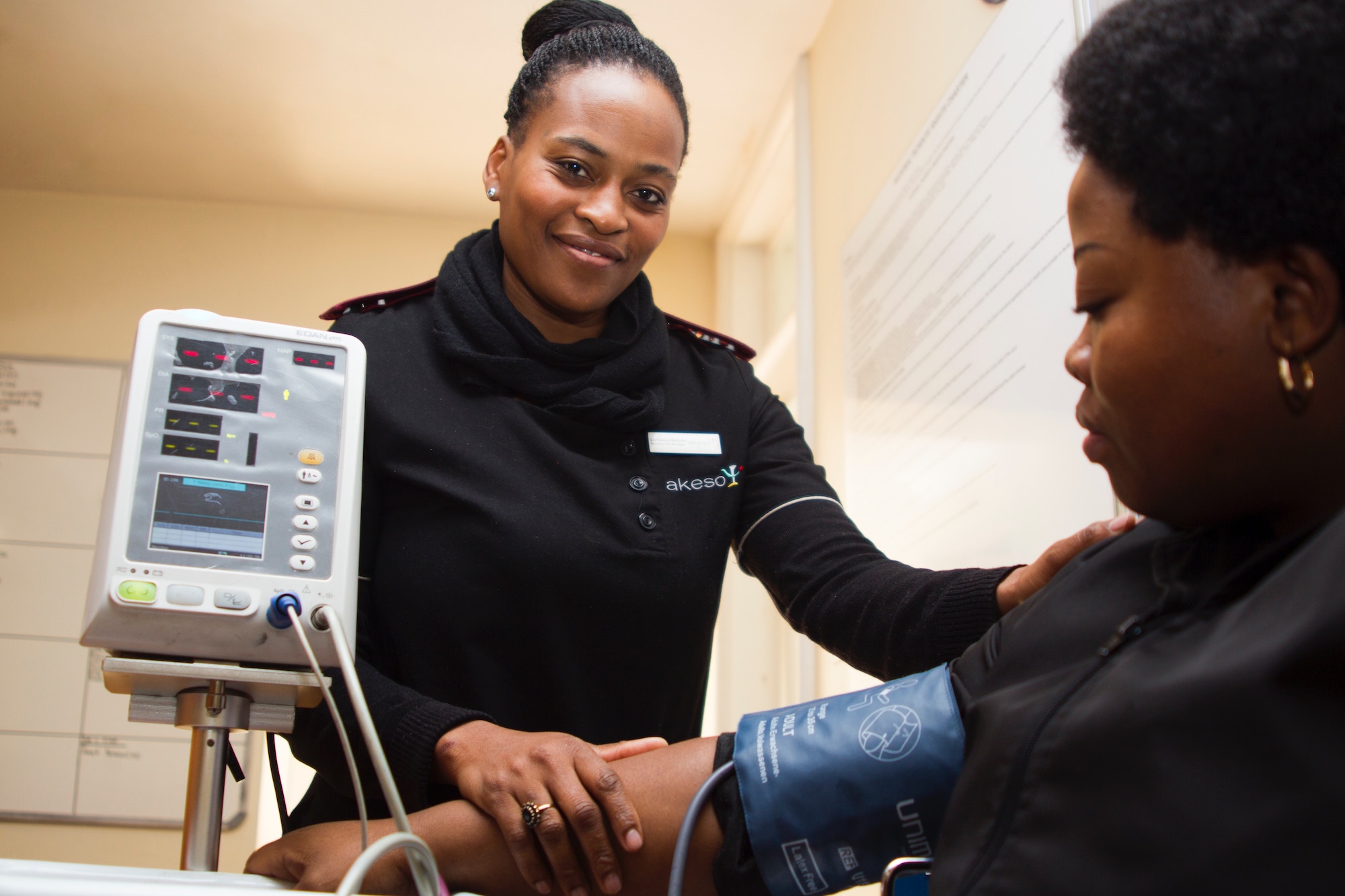 Black nurse in scrubs with her hair up in a bun takes vital signs of a black patient who is sitting in a chair and looking at the blood pressure cuff on her arm