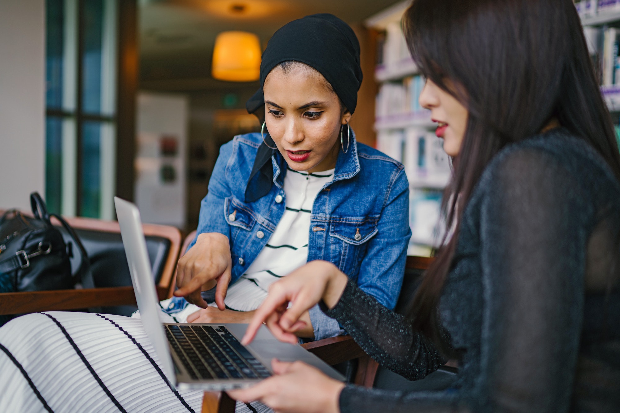 Girl wearing a hijab and long striped dress points to a laptop screen while a woman in a sheer black long-sleeve top points at the same screen