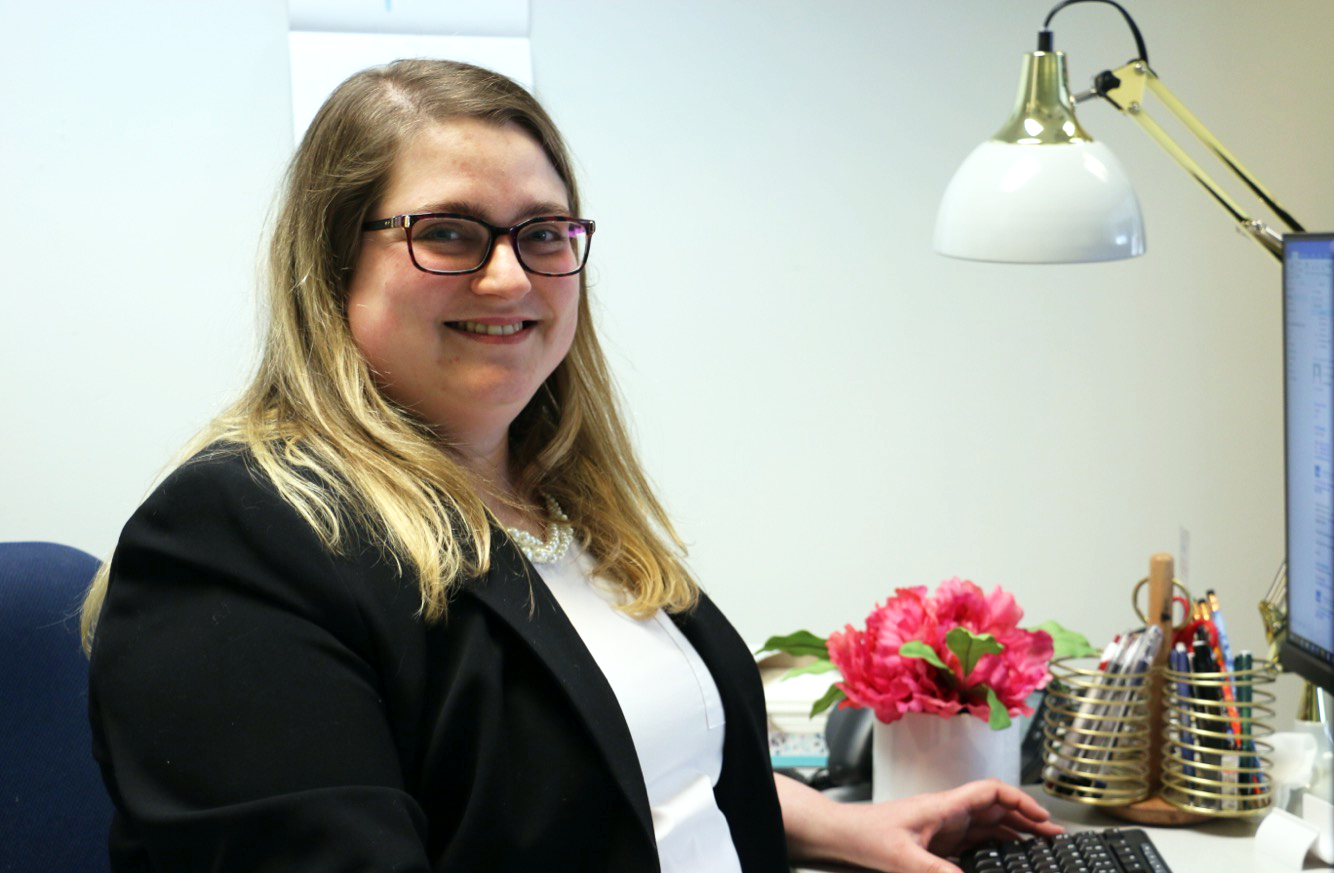 Blonde haired woman in glasses wearing a white tee shirt and black blazer sitting at her desk and smiling