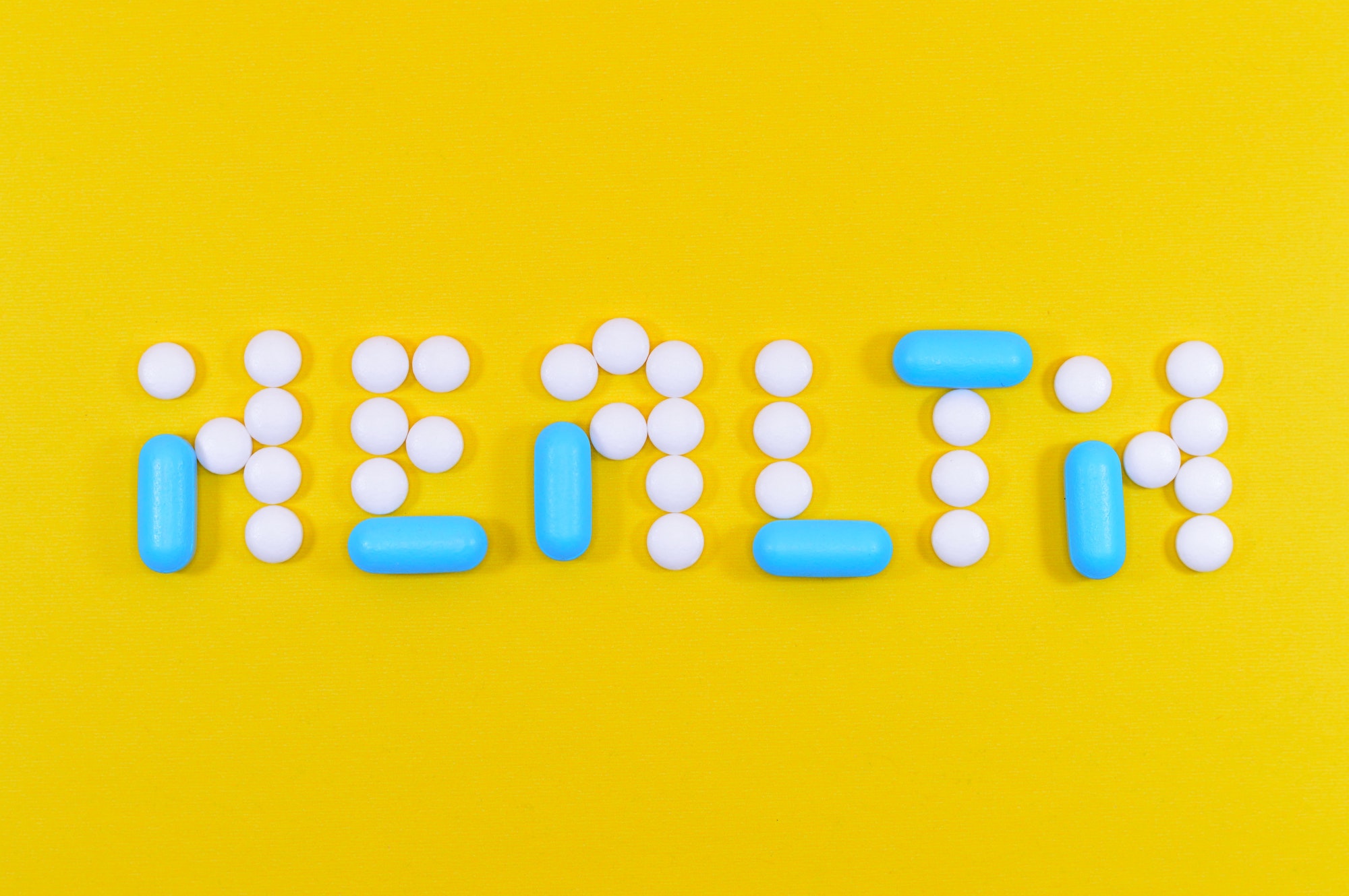 White circular pills and small blue capsule pills are arranged on a canary yellow background and spell the word HEALTH