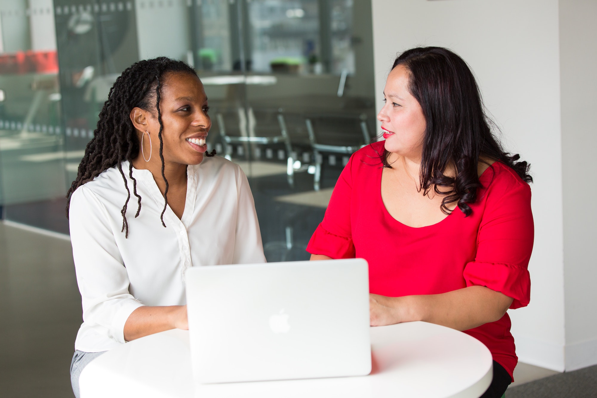 Black woman in a white button down shirt smiling and talking to an Asian woman in a red dress in an office setting