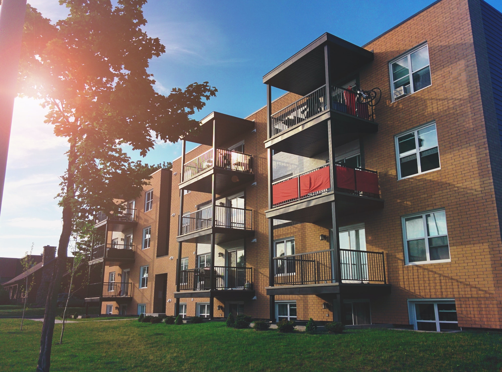 Sunny image of brick apartment building with trees in the forefront and luscious green grass