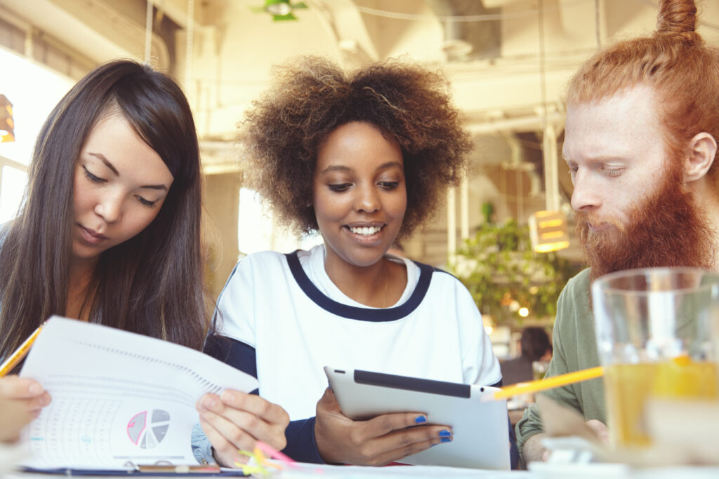 International team work. Three people working together at cafe: Asian woman studying financial report on paper while her African colleague showing new business plan to redhead man using touch pad